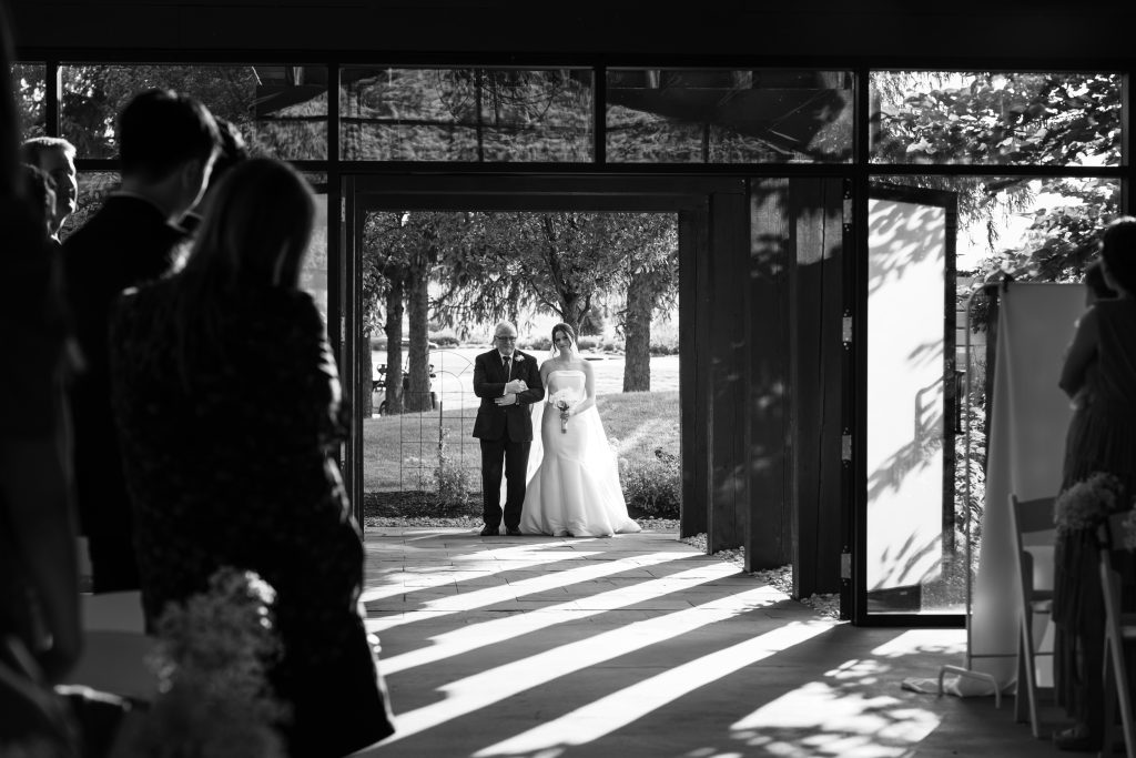 A bride in a white dress walks down the aisle with a man, likely her father, at an indoor wedding ceremony. Guests stand on either side as sunlight streams through large windows—captured beautifully in timeless documentary photography.