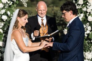A bride in a white dress places a ring on the groom's finger during an outdoor Toronto wedding ceremony. An officiant in a suit stands behind them, holding a book and microphone, with floral arrangements creating an editorial backdrop.