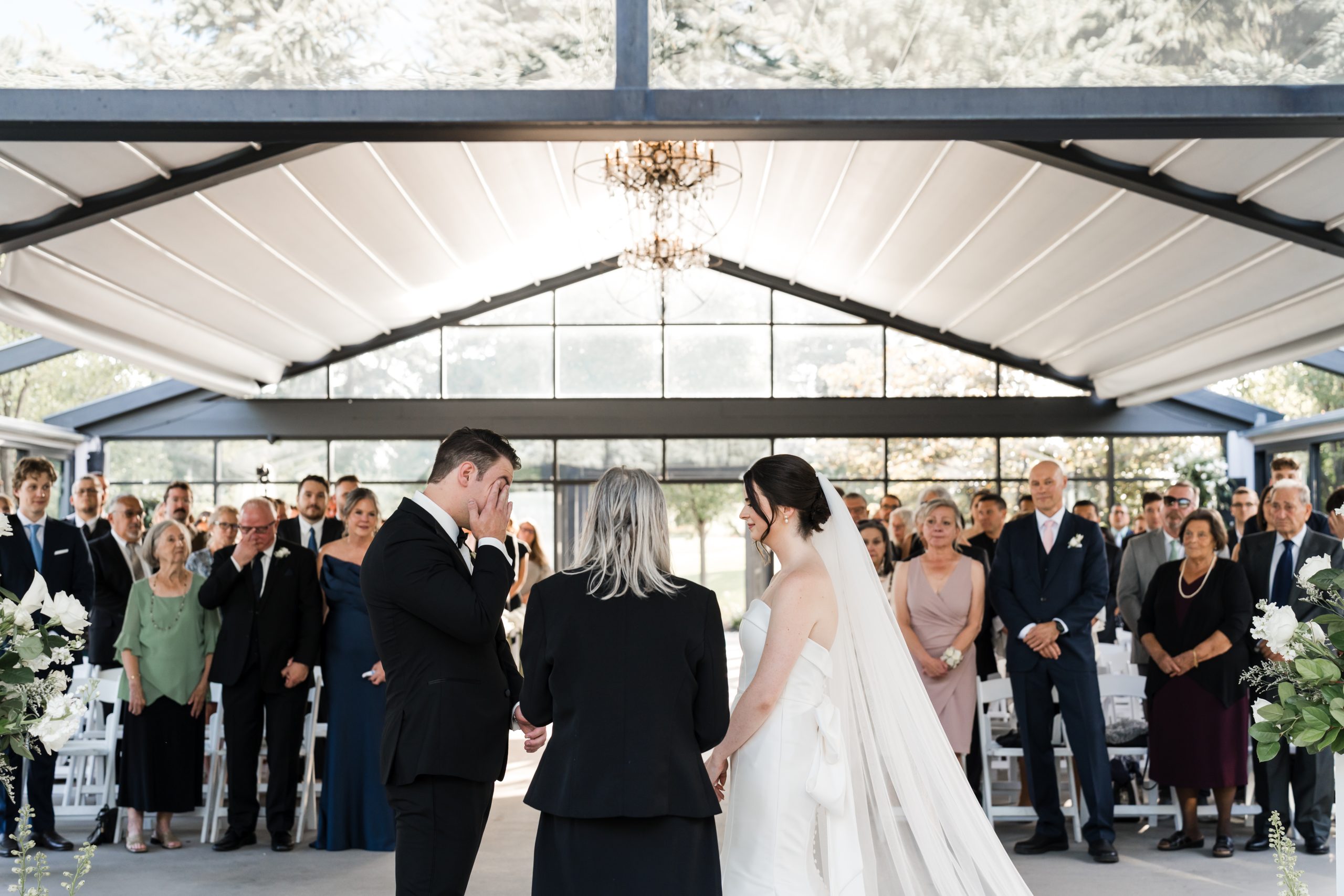 A bride and groom stand facing each other at an indoor wedding ceremony, holding hands. The officiant stands between them as guests watch attentively. Sunlight filters through large windows, creating a beautiful moment of editorial wedding photography.