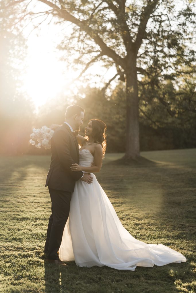 A bride and groom embrace outdoors on a grassy lawn at sunset, with sunlight streaming through the trees behind them. The bride wears a white gown and holds a bouquet, while the groom wears a dark suit.