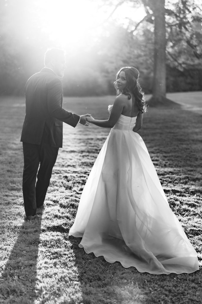 A bride and groom holding hands walk across a grassy lawn in sunlight. The bride smiles back while wearing a flowing gown; the groom wears a suit. The scene is backlit, casting long shadows. Black and white photo.