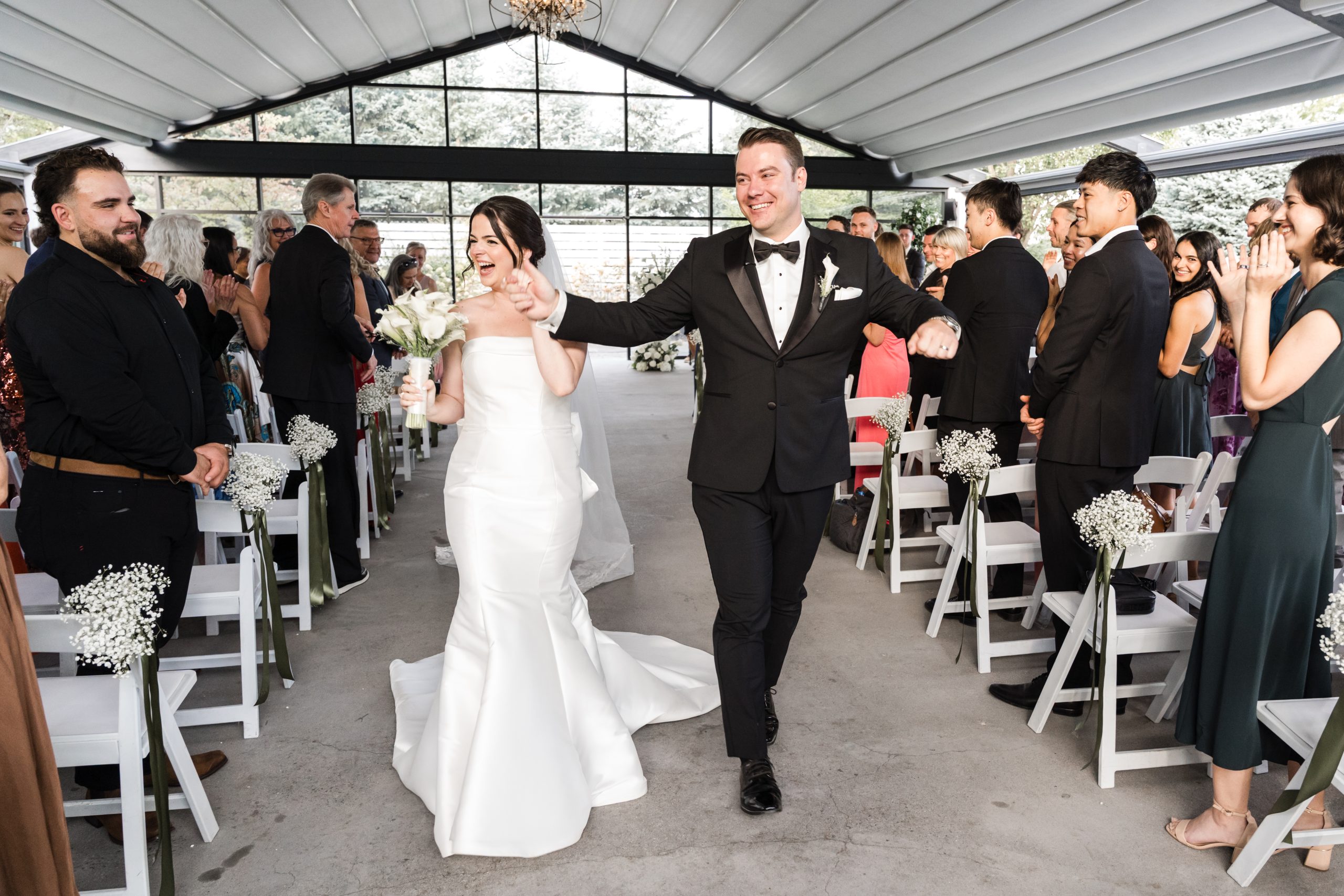 A newlywed couple, captured by a talented wedding photographer, walk joyfully down the aisle together in an indoor venue as guests stand and smile on either side.