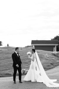 A bride in a long white gown and veil holds a bouquet, smiling at a groom in a black suit as they stand outside on a pathway with grass and a building in the background, captured by a talented Toronto photographer.