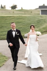 A bride in a strapless white gown and a groom in a black tuxedo hold hands and smile as they walk outdoors on a paved path, captured by a Toronto wedding photographer, with green grass and a building in the background.