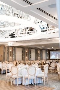 Elegant banquet hall with round tables set for an event, surrounded by light-colored chairs. Modern chandeliers hang from the ceiling, while a projection screen displays a black and white editorial photography image in the background.