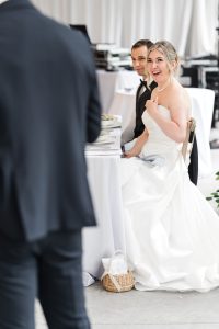 A bride in a white dress sits next to a groom at a table, smiling and looking at someone in front of them. The photography captures an elegant Toronto wedding reception with white tablecloths and beautiful decorations.