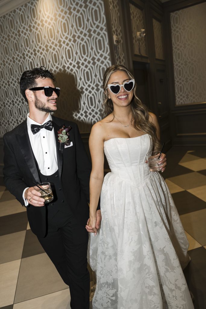 A smiling bride and groom wearing sunglasses hold hands at an indoor wedding venue. The bride is in a strapless white dress with heart-shaped glasses, and the groom is in a black tuxedo with dark sunglasses. Both hold drinks. Captured by an editorial wedding photographer in Toronto
