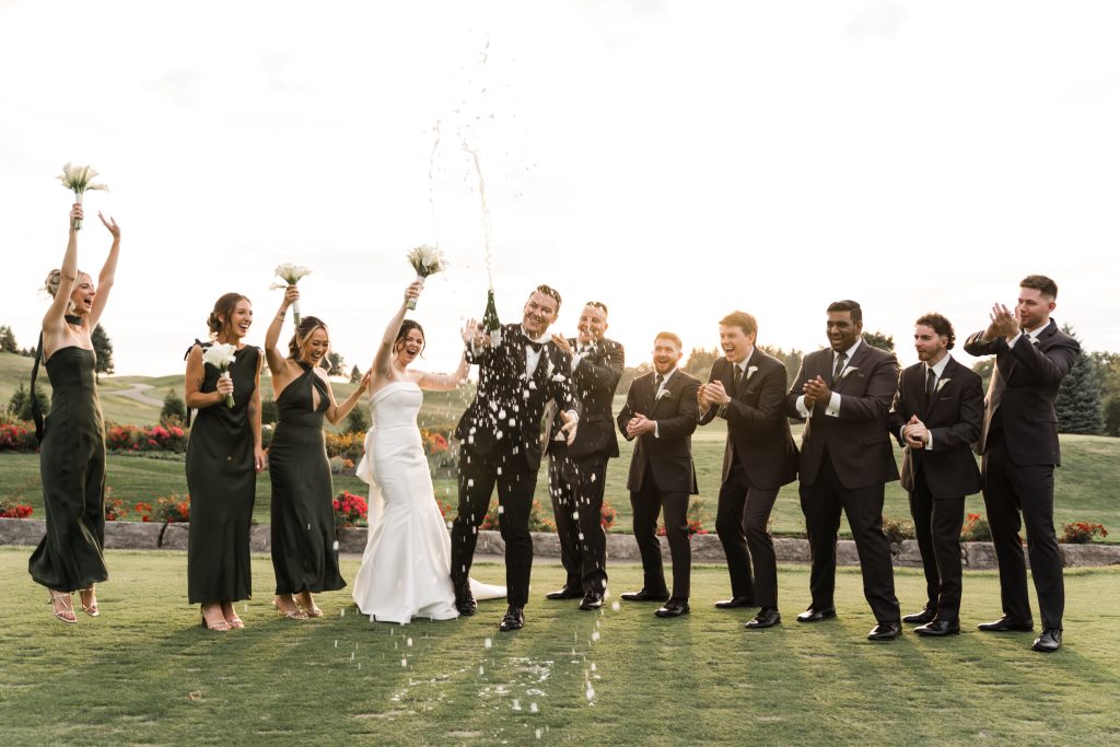 A wedding party celebrates outdoors as the groom pops a champagne bottle, spraying champagne. Bridesmaids in green dresses and groomsmen in black suits cheer and smile on a sunny day with flowers in the background.