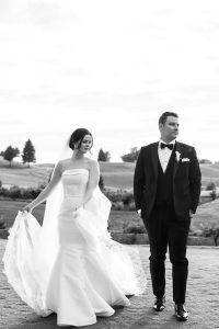 A bride in a strapless white gown and veil walks beside a groom in a tuxedo. Captured in black and white, this wedding photography scene unfolds outdoors on grass, both looking away as the bride holds her dress.