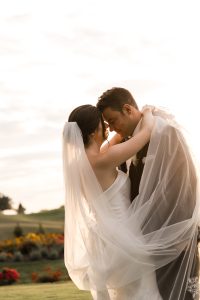 A bride and groom embrace outdoors at sunset, touching foreheads and smiling softly. Captured by a wedding photographer, the bride’s veil and dress glow amid colorful flowers and greenery in this editorial-style moment.