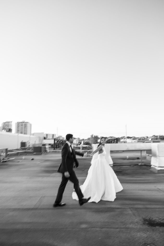 A black-and-white photo of a bride and groom walking together on a rooftop, holding hands. The bride wears a long white dress and veil. City buildings and rooftops are seen in the background under a wide sky.