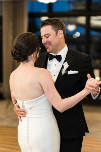 A bride and groom, dressed in a white strapless gown and black tuxedo, smile and dance closely together indoors during their wedding reception in Toronto, capturing a beautiful documentary moment.