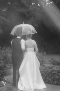 A bride in a white gown and a groom in a vest walk arm in arm under a clear umbrella along a garden path, with lush greenery and soft light giving this wedding scene an elegant, editorial feel.