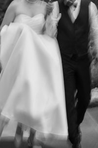 A blurred black-and-white photography captures a bride in a strapless gown and pearl necklace walking hand-in-hand with her groom in a tuxedo, moving together in editorial elegance.