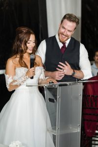 A bride in an off-shoulder white gown with lace sleeves smiles beside her groom at a clear podium, both radiating joy during a wedding speech—captured perfectly by the documentary photographer.
