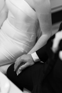 A black and white photo shows a couple sitting close together. The woman in a strapless dress gently holds hands with a man in a suit, their hands resting on his leg—an editorial-style Toronto wedding moment, their faces out of frame.