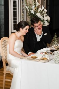 A bride and groom sit side by side at their Toronto wedding reception table, smiling and laughing. The documentary-style photo captures wine glasses, cheese, and crackers on the table in front of them.