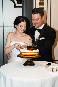 A bride and groom in formal attire smile as they cut a fruit-topped cake together at their wedding reception, as a photographer captures the moment. The cake sits on a stand on a round table with a white tablecloth.