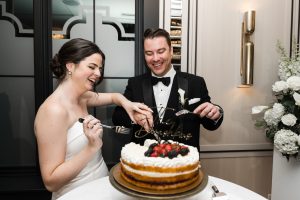 A bride and groom in formal attire laugh as they cut and serve a tiered wedding cake, topped with berries and a gold sign, beside a floral arrangement in an elegant room—a perfect moment for any Toronto photographer’s editorial portfolio.