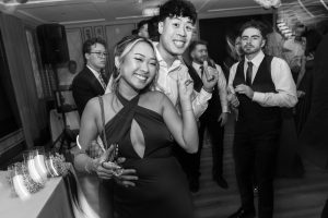 A smiling couple poses playfully on a crowded dance floor at a formal event, surrounded by other well-dressed guests enjoying the celebration, capturing a lively moment in documentary photography.