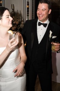 A smiling bride in a white dress and groom in a black tuxedo laugh together while holding drinks at their Toronto wedding reception, captured perfectly by the photographer.