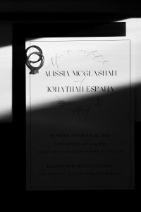 A wedding invitation partly in shadow, with two rings intertwined on the top left corner. The editorial-style text features the names Alissia McGlashan and Johathan Espaha, detailing the wedding event at Eglington West Gallery.