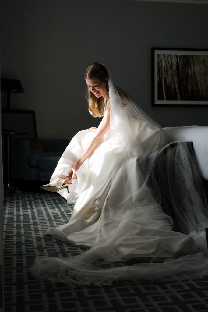 A bride in a white wedding dress and veil sits on the edge of a bed, smiling as she puts on her shoe. Sunlight streams in, highlighting her dress in this documentary photography moment. A chair, lamp, and framed picture sit in the background.