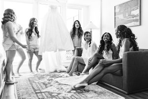 Six women in matching pajamas smile and laugh together in a bright room as a wedding dress hangs behind them. Captured by a photographer, this candid moment creates a joyful, documentary-style atmosphere among friends.