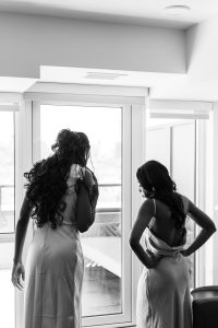 Two women in satin dresses stand by a large window, looking outside. Captured in black and white, this photography scene shows one woman leaning forward while the other stands with hands on her hips.