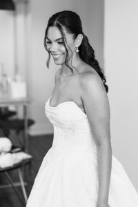 A smiling woman in a strapless wedding dress with her hair styled in loose waves stands indoors in a softly lit room. This black and white photograph captures an editorial elegance through the photographer’s artistic vision.