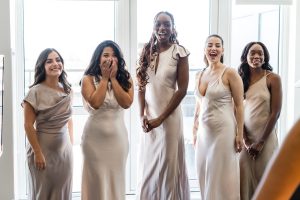 Five women in matching light beige dresses stand indoors, smiling and laughing in front of large windows with bright natural light streaming in. The scene feels editorial, as one woman covers her mouth in surprise or excitement.