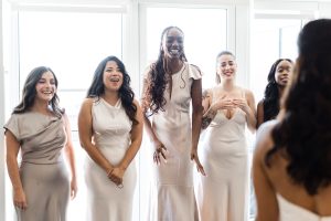 Five women in light-colored dresses stand together indoors, smiling and laughing toward the wedding photographer in the foreground. Bright light pours through large windows behind them, capturing a joyful moment in this beautiful wedding photography scene.