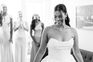 A bride in a strapless gown smiles and looks down, while three bridesmaids in light dresses stand in the background, smiling and applauding—a perfect moment captured by a wedding photographer. The scene is bright and joyful.