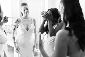 A group of women in light dresses share an emotional moment; one wipes tears from her face while another looks on sympathetically, captured by a photographer in a bright room with soft natural light.