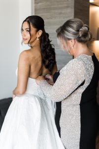 A bride in a white strapless gown looks over her shoulder as a woman in a sparkly black and silver dress helps fasten the back, captured by a Toronto documentary photographer.