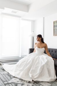A woman in a strapless white wedding dress sits on a dark gray couch in a bright, modern room with large windows, looking to the side and smiling slightly as the photographer captures this elegant moment.