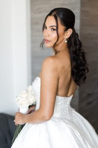 A bride in a strapless white wedding dress holds a bouquet of white roses and looks over her shoulder. Captured in elegant wedding photography, she has long, dark, wavy hair, pearl earrings, and a calm expression.