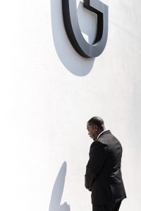 A man in a black suit stands against a white wall with a large black letter G mounted above him. The sunlight casts his shadow and the letter’s shadow on the wall, creating a striking image suited for any Toronto documentary photographer.