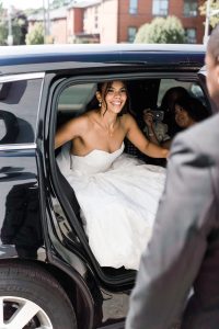 A smiling bride in a white strapless gown steps out of the backseat of a car, assisted by a person in a suit. Behind her, someone holds a camera, capturing this documentary moment. Trees and Toronto buildings fill the background.