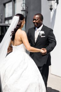 A bride in a strapless white gown and veil holds hands and smiles with a man in a black suit and white boutonniere, standing outdoors in bright sunlight—captured by a Toronto editorial photographer.
