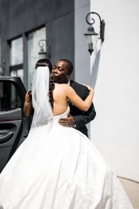 A bride in a white dress and veil embraces and shares a kiss on the cheek with a man in a black suit outdoors, near a building and car, in bright sunlight—capturing a candid moment of wedding documentary photography.