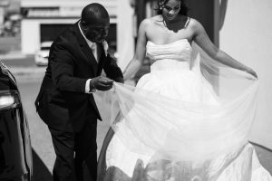 A man in a suit helps a woman in a strapless wedding dress arrange her veil or train outdoors on a sunny day, captured in black and white documentary photography.