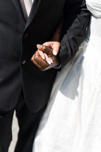 A close-up documentary photograph of a couple holding hands, one in a black suit and the other in a white dress, highlighting their intertwined fingers and contrasting skin tones.