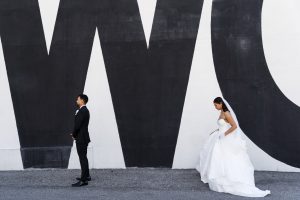A bride in a white dress walks toward a groom in a black suit, standing apart in front of a large wall painted with bold black and white letters—captured with striking artistry by the photographer.