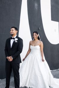 A bride in a strapless white gown smiles behind her groom in a black tuxedo with a white boutonniere. Captured by their wedding photographer, the couple poses against a striking black-and-white wall in Toronto.