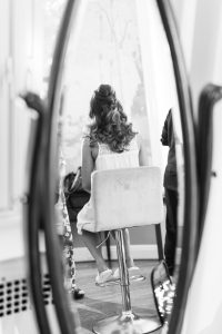A woman with curled hair sits on a high chair, seen from behind in a mirror reflection, wearing a light dress and sandals in a softly lit room. The black and white photograph captures timeless beauty through the lens of a Toronto photographer.