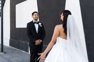 A groom in a black tuxedo smiles at a bride in a white wedding dress and veil, standing with open arms outdoors against a black and white geometric wall—captured by a Toronto photographer with an editorial flair.