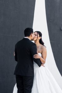 A bride and groom share a kiss in front of a modern black and white wall. Captured by their wedding photographer, the couple embrace closely—her in a strapless gown, him in a black suit—creating a romantic documentary moment.