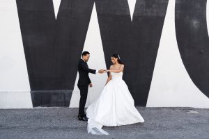 A bride in a white gown and a groom in a black suit stand in front of a bold black and white wall, holding hands and smiling at each other—a perfect moment of editorial photography.