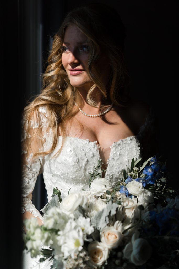 A bride with long wavy blonde hair in a white lace dress holds a bouquet of white and blue flowers. Soft sunlight illuminates her face as she gazes to the side, wearing a pearl necklace—captured by a Toronto wedding photographer.